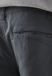 Pantalon gris foncé en tissu lisse, doté d'une poche arrière avec une étiquette de logo discrète et des détails de couture soignés.