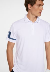 Vitt polo skjorta med uppvikt krage, med en marinblå rand på ärmarna och en liten logotyp på kragen. Slät tygtextur.