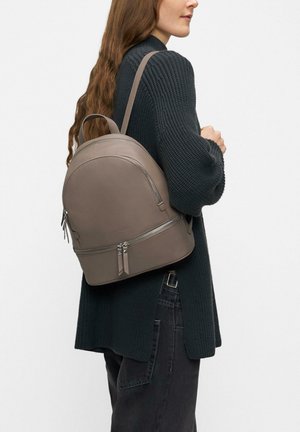 Frau in schwarzem Strickpullover trägt taupefarbenen Leder-Rucksack mit zwei Reißverschlussfächern über einer Schulter.