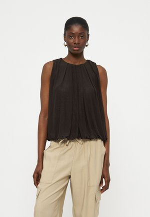 Γυναίκα που φοράει μαύρη αμάνικη μπλούζα με πιέτες και μπεζ παντελόνι cargo, στέκεται μπροστά σε λευκό φόντο.