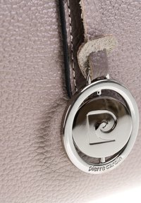 Charm en argent en forme de cercle avec le logo Pierre Cardin, attaché à un cuir taupe texturé avec des coutures noires.