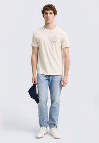 Beige t-shirt med korta ärmar och cirkulärt märke på vänster bröst, kombinerad med ljusblå jeans och vita sneakers, håller en marinblå keps i vänster hand.