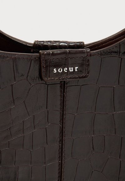 Υφή από τεχνητό δέρμα κροκό σε σκούρο καφέ χρώμα με ραμμένη ετικέτα που δείχνει το όνομα της μάρκας «soeur» με λευκά γράμματα στη λαβή της τσάντας.