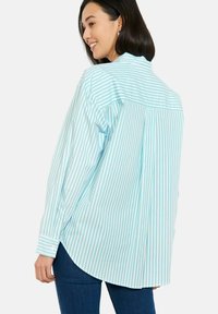 Tumejuustega naine naeratab, kandes helesinise ja valge vertikaaltriibulise pika varrukaga särki ja tumedasiniseid teksaseid, ees veidi pööratud.