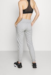 Person som bär grå sweatpants, svart sport-bh och svarta sneakers, står med händerna i fickorna mot en enhetlig bakgrund.