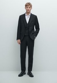 Schwarzer, maßgeschneiderter Anzug mit einem einreihigen Jacket, passender Weste und schmal geschnittenen Hosen. Ein weißes Hemd und schwarze Lederschuhe runden den Look ab.