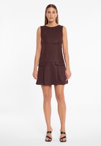 Bordeauxfarbendes ärmelloses Kleid mit gerippter Textur, das zwei Taschen auf der Vorderseite und einen ausgestellten Rock hat. Tragen mit schwarzen Riemchensandalen.