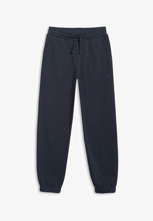 Jeune fille aux longs cheveux bruns portant un sweat à capuche bleu marine zippé, un jogging assorti, un crop top blanc et des baskets noires et blanches sur un fond neutre.