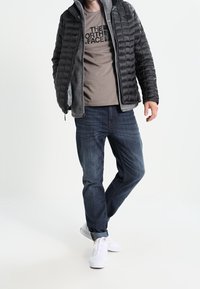 Czarna kurtka puchowa o pikowanym wzorze na szarym podkoszulku, zestawiona z ciemnymi dżinsami i białymi trampkami. Swobodny strój, nowoczesny design.