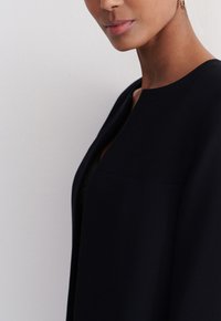 Schwarze, strukturierte Jacke mit runder Ausschnitt, glatter Textur und minimalistischer Gestaltung. Der Stoff wirkt dick und hochwertig.