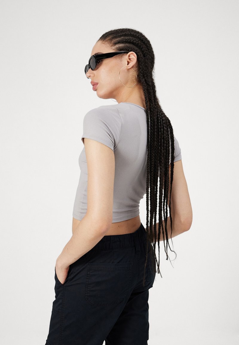 Graues, kürzes T-Shirt, mit kurzen Ärmeln, taillierte Passform. Schwarze Sonnenbrille, goldene Creolen. Schwarze Hose mit elastischem Bund, seitlich sichtbare Tasche.