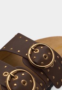 Κοντινό πλάνο σκούρων καφέ λουριών σανδαλιού με στρογγυλά χρυσά αγκράφες και διασκορπισμένες μικρές χρυσές καρφίτσες πάνω σε δερμάτινο υλικό.