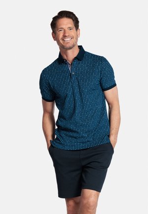 Marineblauwe polo met een geometrisch patroon, een sluiting met twee knopen en een contrasterende donkere kraag. Gecombineerd met zwarte shorts, de stof lijkt zacht.