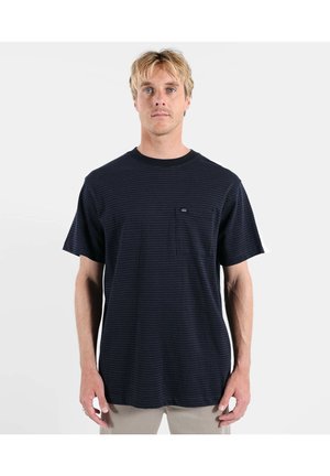 Un homme blond se tient face à l'avant, portant une chemise noire texturée à manches courtes avec une petite poche sur la poitrine et un pantalon beige.