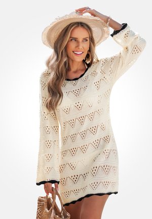 Vestito di maglia color crema con motivi a zigzag traforati e maniche lunghe, rifinito in nero. Il modello tiene in mano una borsa intrecciata e indossa un cappello a tesa larga.