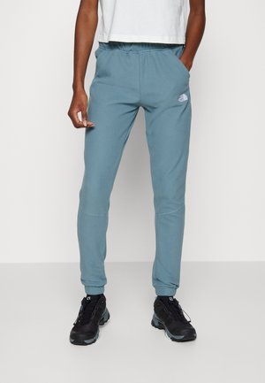 Persoon draagt blauwe joggingbroek met een klein logo op de linker dij, wit shirt en zwarte sportschoenen, staat met één hand in de zak.