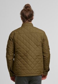 Oliivivihreä tikattu takki, jossa on pystykaulus, nappikiinnitys ja teksturoitu kangas, jossa on timanttikuviot selkäpuolella.