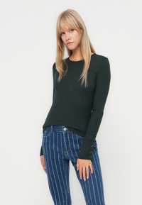 Mørkegrønn ribbestrikket genser med lange ermer, rund hals og knappdetaljer på ermene, kombinert med blå stribede jeans.