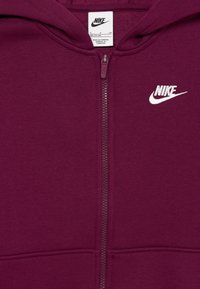 Bordo zip-up džemperis pagamintas iš minkštos medžiagos. Yra baltas Nike logotipas ant krūtinės, lygus užtrauktukas ir kengūros kišenė priekyje.