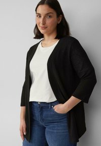 Zwarte cardigan met driekwart mouwen, open voorkant, gemaakt van gestructureerd materiaal, gedragen over een wit T-shirt en blauwe spijkerbroek.