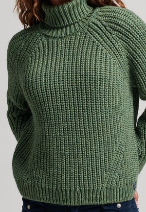 Frau trägt einen grob gestrickten olivgrünen Rollkragenpullover mit langen Ärmeln und geripptem Muster.