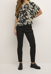 Bloemige blouse met een zwart en crème patroon, losse pasvorm, korte mouwen, gecombineerd met slanke zwarte broek en zwarte schoenen, staand voor een neutrale achtergrond.