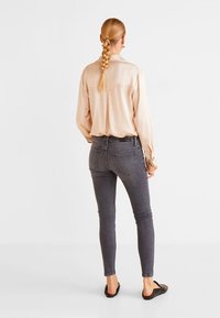 Camisa de manga larga beige claro combinada con jeans skinny gris oscuro y zapatos planos negros, vista desde atrás. Tejido suave, con detalles mínimos.