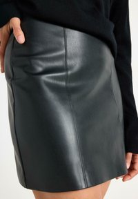 Sort læder mini skirt med glat tekstur, der har vertikale sømme og en tætsiddende silhuet, holdt af en hånd på en neutral baggrund.