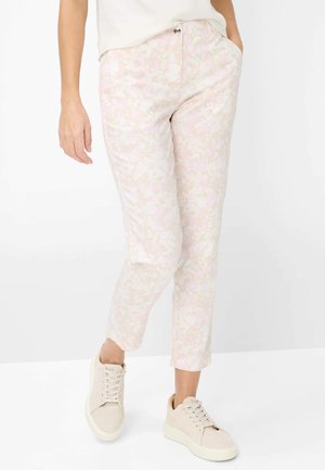 Persona che indossa pantaloni corti floreali color rosa chiaro con tasche frontali e scarpe da ginnastica beige con lacci, in piedi contro uno sfondo bianco.