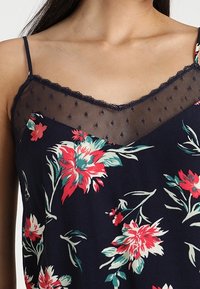 Top in stile floreale di colore navy, con fiori rossi e turchesi. Bordo in pizzo con rete a pois sul colletto. Spalline sottili regolabili.