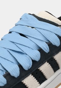 Dark blue sneaker featuring textured fabric, light blue flat laces, and a beige stripe design on the side, with visible stitching details.