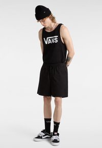 Musta toppi, jossa valkoinen "VANS"-logo, mustat shortsit, mustat sukat, joissa on valkoinen logo, sekä mustat ja valkoiset lenkkarit. Mallilla on päässään musta pipo.