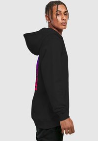 Sort hættejakke med et farverigt grafisk design i lilla og pink på ryggen, med en kengurulomme og snoretræk i hætten.