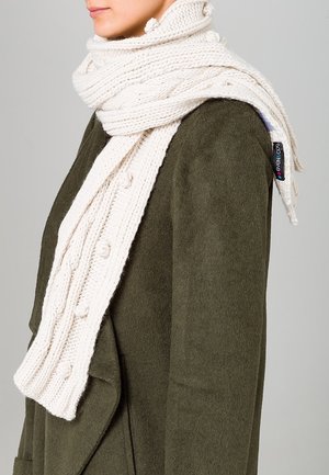 Personne portant une épaisse écharpe tricotée crème avec des détails à pompons sur un manteau en laine vert foncé, tourné vers la gauche sur un fond uni.