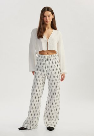 Afskåret hvid bluse med frontknude og lange ærmer, parret med løstsiddende hvide bukser med et sort blomstermønster. Sorte slip-on sko.