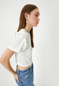 Hvid cropped top med tekstureret stof og voluminøse korte ærmer, parret med blå denim jeans. Model vist i profil.