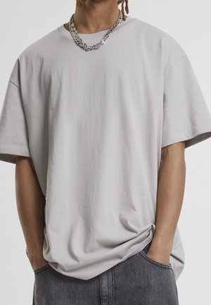 Person iført en løs lysegrå T-shirt, mørkegrå jeans, sølvkæde, med hænderne i lommerne mod en ensfarvet baggrund.