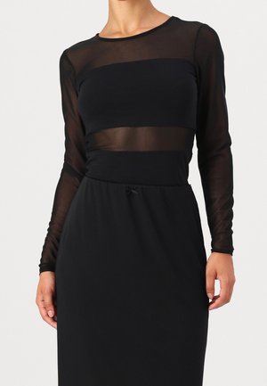 Vrouw draagt een zwarte jurk met lange mouwen en doorzichtige meshpanelen over de bovenborst, mouwen en taille, staand tegen een effen achtergrond.