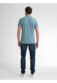 Camisa polo de color azul claro con mangas cortas, combinada con jeans azul oscuro y zapatillas blancas, vista desde la parte de atrás sobre un fondo liso.