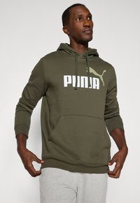 Mand justerer sin olivengrønne Puma hoodie med hvidt logo og grå sweatpants foran en lys ensfarvet baggrund.