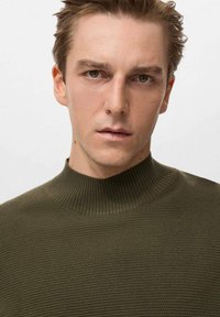 Maglione a collo alto a coste di colore verde oliva con un colletto alto e un tessuto morbido e texturizzato. Inquadratura ravvicinata della parte superiore del corpo.