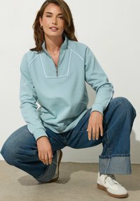 Felpa azzurra chiara con zip, con piping bianco sul colletto e sulle spalle, abbinata a jeans blu larghi e corti e scarpe bianche.