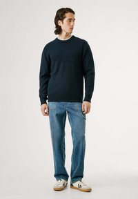 Jūrų pajėgų megztinis su raštuota apykakle, derinamas su šviesiai mėlynomis džinsinėmis kelnėmis. Sportbačiai su baltais ir juodais akcentais. Kasdienis stilius.