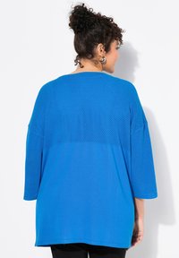 Blå mesh- och solid tröja med trekvartslånga ärmar och rundad halsringning. Har en texturerad övre del och en slät nedre del.