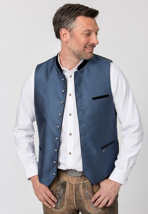 Mann trägt weißes Hemd, blaue gemusterte Weste und braune bestickte Lederhosen, schaut nach links mit den Händen auf den Hüften.