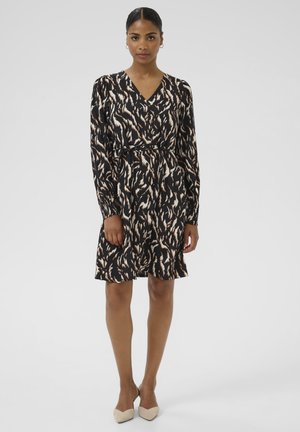 Femme debout portant une robe longue à manches longues en imprimé abstrait noir, beige et marron avec un décolleté en V et des chaussures à talons beige.