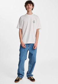 Biała koszulka z krótkim rękawem z graficznym wzorem na lewej piersi, w połączeniu z jasnoniebieskimi, luźnymi jeansami i brązowymi trampkami.