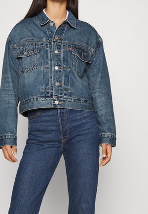 Femme portant une veste en denim surdimensionnée raccourcie avec fermeture à boutons et un jean bleu taille haute sur fond uni.