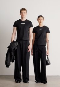 T-shirts pretas de algodão com logótipo branco "TOMMY", combinadas com calças pretas largas. A modelo feminina segura uma pequena mala, ambos usam calçado preto.