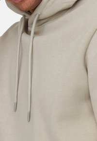 Lys beige hoodie med snore, der har en glat tekstur og et afslappet design. Nærbillede af stoffet og syningerne.